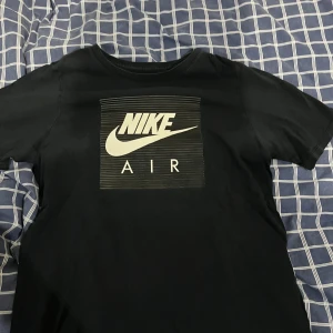 Svart Nike Air t-shirt med tryck - Svart t-shirt från Nike med stort vitt Nike Air-tryck på bröstet och tunna horisontella linjer i bakgrunden. Klassisk rund hals och korta ärmar. Tillverkad i mjuk bomull för skön känsla och snygg passform.