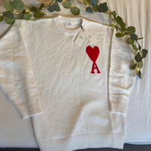 Vit stickad tröja från Ami Paris - Vit stickad tröja från Ami Paris med röd hjärtlogga och bokstaven A på bröstet. Tröjan har rund halsringning, ribbade muddar och är tillverkad i Portugal. Perfekt för dig som gillar stilrena och trendiga plagg med en twist.
