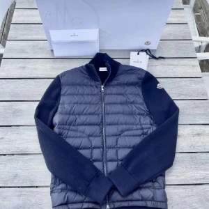 Moncler cardigan - Skriv om du har frågor! Snygg mörkblå cardigan från Moncler med quiltad framsida och stickade ärmar. Västen har dragkedja framtill och en liten Moncler-logga på ena ärmen. Perfekt för dig som gillar exklusiva märken och stilren design.