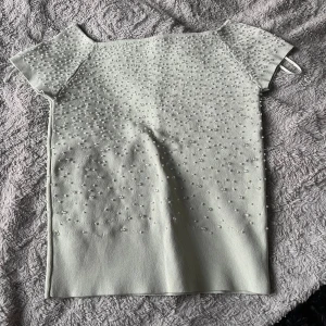 Vit topp med pärldetaljer - Supersöt vit topp med korta ärmar och massor av små pärlor över hela framsidan och axlarna. Toppens raka modell ger en clean look och pärlorna gör den extra speciell. Perfekt för dig som vill sticka ut lite extra. Mint grön färg 