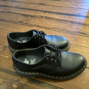 Svarta Dr. Martens vegan skor - Säljer dessa klassiska svarta Dr. Martens i storlek 38, lite midre i storleken dock. Skorna är tillverkade i syntetmaterial och har den ikoniska gula sömmen runt sulan. Luftdämpad sula för extra komfort och slitstyrka. Passar dig som vill ha en stilren och tidlös look.
