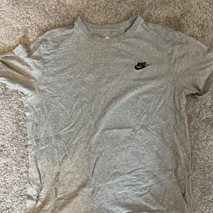 Grå Nike t-shirt i bomull - Snygg grå t-shirt från Nike med klassisk svart logga på bröstet. T-shirten har rund halsringning och korta ärmar. Tillverkad i mjuk bomull för en skön känsla hela dagen.