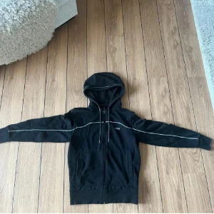 Svart hoodie från BOSS med dragkedja (sällsynt) - Svart hoodie från BOSS med vit piping längs ärmar och axlar, samt logga på bröstet. Tröjan har huva med snörning, två fickor fram och hel dragkedja. Materialet är mjuk bomull och passformen är normal. Perfekt för chill eller streetwear.