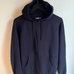 Svart hoodie från Tommy Hilfiger - En stilren svart hoodie från Tommy Hilfiger med huva och snörning. Tröjan har en klassisk passform, känguruficka framtill och är tillverkad i mjuk bomull. Perfekt för en avslappnad look och enkel att matcha med jeans eller joggers.