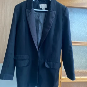 Svart kavaj från H&M, storlek 36 - Klassisk svart kavaj från H&M i storlek 36. Kavajen har släta slag i satin, två fickor med lock framtill och en enkel knäppning. Fodrad insida.