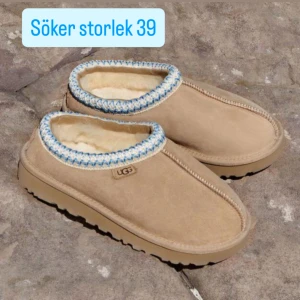 SÖKER Beige UGG slip-ins med ullfoder - Söker i storlek 39. Mysiga slip-in skor från UGG i beige mocka med fluffigt ullfoder. Skorna har en dekorativ kant i blått och vitt runt öppningen och en rejäl platt sula. Perfekta för kalla dagar när du vill ha det extra bekvämt.