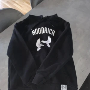 Svart Hoodie från Hoodrich med tryck - Svart hoodie från Hoodrich med vit text och motiv av duvor på både fram- och baksidan. Baksidan har även blå text 'From Nothing To Something' och vit text 'Product of the Streets Est. 2014'. Tröjan har huva med snörning och ribbade muddar.
