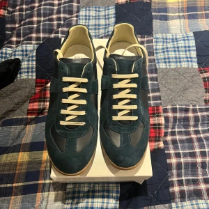 Maison Margiela sneakers blå mocka - Snygga sneakers från Maison Margiela i mörkblå mocka och skinn med beige snörning och gummisula. Klassisk siluett med rund tå och diskret vit detalj bak på hälen. Perfekta för dig som gillar stilrena och exklusiva sneakers.