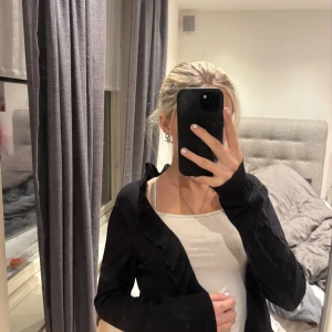 Svart volangkofta från H&M - Svart kofta från H&M i storlek S med snygga volangdetaljer längs hela kanten och nederdelen. Koftan är långärmad och har en ribbad struktur vid volangerna. Perfekt att slänga över en topp för en chill och stilren look.