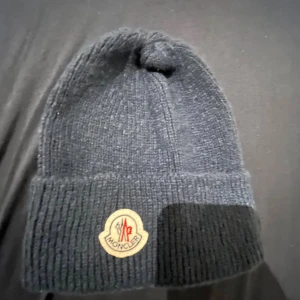 Mörkgrå stickad Moncler mössa - Snygg mörkgrå ribbstickad mössa från Moncler med klassisk uppvikt kant och ikonisk logotyp framtill. Perfekt för kalla dagar och ger en clean streetstyle-vibe. Materialet är mjukt och värmande, och mössan har en enkel, tidlös design.
