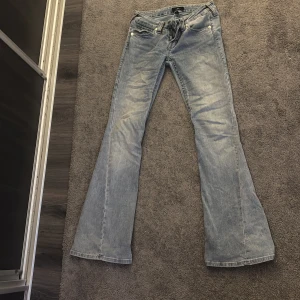 True Religion Joey Low Rise Flare Jeans - Säljer ett par ljusblå jeans från True Religion, modell Joey Low Rise Flare. De har låg midja, utställda ben och klassiska fickor med knappar bak. Jeansen är tillverkade i denim och har snygga slitningar för en cool vintagekänsla.
