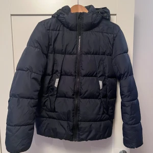 Mörkblå pufferjacka från Tom Tailor - Säljer en mörkblå pufferjacka från Tom Tailor Denim med huva och dragkedja framtill. Jackan har två fickor med reflexdetaljer och är quiltad för extra värme. Perfekt för kalla dagar och har en clean, modern look som funkar till det mesta. Mycket fint skick. 