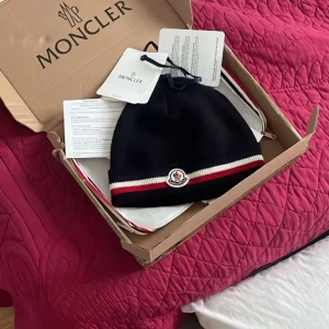 Svart Moncler mössa med röd/vit kant - Snygg svart mössa från Moncler med klassisk logga framtill och röd/vit rand längs kanten. Perfekt accessoar för att hålla dig varm och stilren under kyliga dagar. Mössan är ribbstickad och har NFC tagg.( endast mössa)