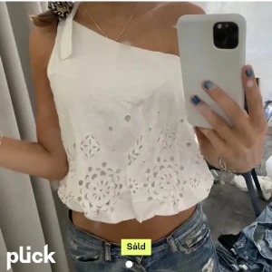 Vit oneshoulder broderad topp - Säljer en vit oneshoulder topp med broderade detaljer och hålmönster. Toppen är croppad och har en rosett på axeln.