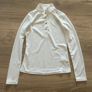 Vit ridtröja - Snygg vit polotröja med lång ärm och silverfärgade knappar framtill. Tröjan har ett broderat logotypmärke på bröstet och är tillverkad i mjukt, stretchigt material som passar perfekt till ridning. Stilren och sportig design, passar perfekt till tävling. 