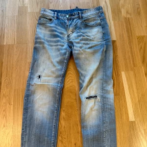 Blå jeans från Dsquared2 - Säljer ett par blå jeans från Dsquared2 med coola slitningar och hål på benen. Jeansen har klassisk femficksdesign, ljus tvätt och Dsquared2-logga på bakfickan. Perfekt för dig som gillar en edgy och avslappnad stil.