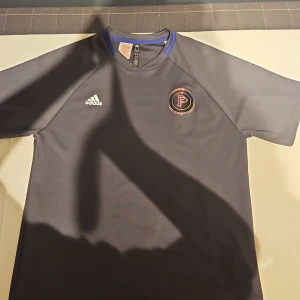 Mörkgrå Adidas fotbollströja L - Snygg mörkgrå fotbollströja från Adidas med korta ärmar och blå detalj i kragen. Framsidan har Adidas-logga och ett färgstarkt P-märke, baksidan har en stor tonad P-grafik. Tillverkad i återvunnet material. Det är tröja av Paul Pogbas kollektion i adidas.