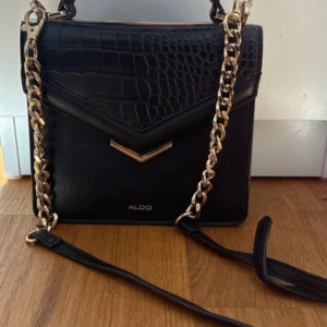Svart axelväska från ALDO med kedja - Snygg svart axelväska från ALDO med guldfärgad kedja och detaljer. Väskan har croco-mönstrad front, stilren form och justerbar axelrem. Insidan har fack med dragkedja för småsaker. Perfekt för dig som vill ha en trendig och praktisk väska.
