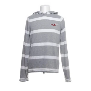 Gråvit randig hoodie från Hollister - Säljer en grå och vit randig hoodie från Hollister med klassisk logga på bröstet. Tröjan har långa ärmar, huva och är tillverkad i mjuk bomull. 
