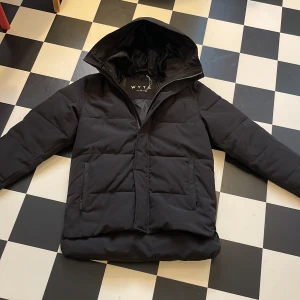 Svart pufferjacka från WYTE med huva - Säljer en svart pufferjacka från WYTE i storlek S. Jackan har en stor huva, två rejäla framfickor och en snygg logga på ärmen. Håller värmen bra till vintern så perfekt för kalla dagar med sin dunfyllda design och dragkedja. Säljes pågrund av felköp, jackan är i nyskick. Storlek S (Stor i storleken, går även som M)