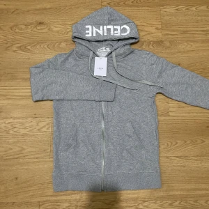 Grå hoodie från Celine med dragkedja - Snygg grå hoodie från Celine med vit logga på huvan, hel dragkedja framtill och praktiska fickor. Tröjan har långa ärmar, snörning vid huvan och är tillverkad i mjuk bomull. Perfekt för chill dagar och streetstyle.