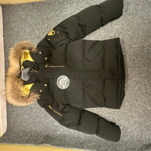 Svart dunjacka Moose Knuckles M - Säljer en svart dunjacka från Moose Knuckles med guldfärgat innerfoder och pälskantad huva. Jackan har flera fickor, guldiga dragkedjor och snygga detaljer på ärmen. Perfekt för kalla dagar när du vill hålla stilen.