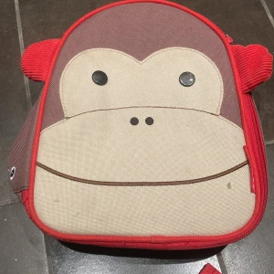 Brun och röd ryggsäck från Skip Hop - Söt ryggsäck från Skip Hop Zoo med apa på framsidan. Väskan är brun och beige med röda detaljer och öron, har justerbara axelband och dragkedja. Tillverkad i slitstark polyester och har ett rymligt huvudfack med innerficka.