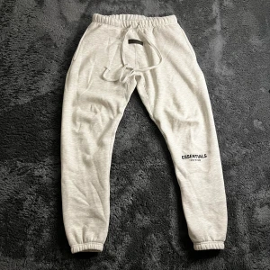 Essentials Fear of God ljusgrå mjukisbyxor - Ljusgrå mjukisbyxor från Essentials Fear of God med elastisk midja, snörning och logga tryckt på benet. Byxorna har muddar vid bensluten och är tillverkade i mjukt bomullsmaterial för maximal komfort. Perfekta för en chill och avslappnad stil.