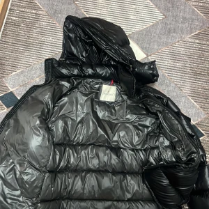 Svart Moncler dunjacka med huva - Säljer en svart dunjacka från Moncler med glansig finish och huva. Jackan har en klassisk Moncler-logga på ärmen, dragkedjor och stora quiltade partier. Perfekt för kalla dagar när du vill hålla stilen och värmen.