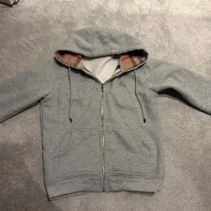 Grå Burberry hoodie med dragkedja - Grå hoodie från Burberry med klassiskt rutigt foder i huvan och längs insidan. Tröjan har huva med snörning, två fickor fram och hel dragkedja. Materialet är mjuk bomull med inslag av elastan för extra komfort. Perfekt för dig som gillar stilrena och lyxiga streetwear-plagg.