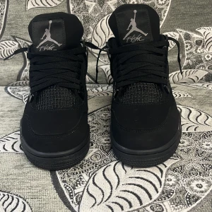 Svarta Nike Jordan 4 black cats sneakers - Säljer ett par svarta Nike Jordan 4 black cats sneakers med klassisk Jumpman-logga på plösen och hälen. Skorna har snörning, meshdetaljer på ovansidan och en robust platt sula. Materialet är en mix av skinn och syntet, vilket ger en cool och sportig vibe.