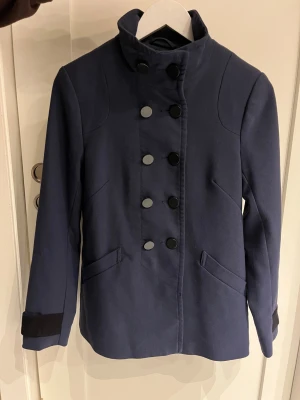 Marinblå kavaj från H&M med knappar - Snygg marinblå kavaj från H&M med dubbelknäppning och hög krage. Materialet är mjukt och är en blandning av polyester och viskos. Perfekt för dig som gillar stilrena och klassiska vibbar. 