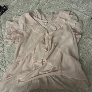 Ljusrosa volangblus med kort ärm - Söt ljusrosa blus i transparent material med volangdetaljer längs ärmar och urringning. Blusen har v-ringning och knyt framtill, samt korta ärmar med volanger för en romantisk look.