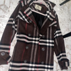Brun rutig trenchcoat från Burberry - Säljer en klassisk trenchcoat från Burberry i brunt med ikoniskt rutigt mönster i svart, vitt och rött. Jackan har dubbelknäppning, bred krage och snygga detaljer vid axlar och ärmslut. Perfekt för dig som vill ha en tidlös och cool look.