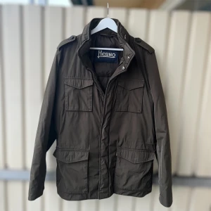 Olivgrön jacka från Herno - Snygg olivgrön parkajacka från Herno med fyra stora fickor framtill, axelklaffar och hög krage. Jackan har dragkedja och tryckknappar, perfekt för dig som gillar stilren och funktionell design. Storlek 54 men sitter mindre än så, mer som en 50 vilket motsvarar en M/L
