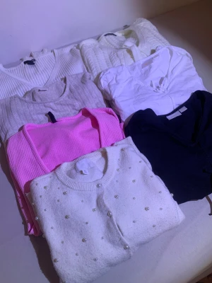 Mix av koftor och tröjor i olika färger - Säljer ett paket med sex tröjor och koftor i olika stilar och färger. Inkluderar en vit ribbad v-ringad tröja, en vit stickad kofta, en grå ribbad tröja, en rosa ribbad tröja, en vit kofta med pärlor från H&M och en svart kofta med snörning framtill. Perfekt för att variera din look!