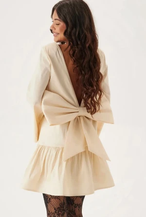 Champagnefärgad kortklänning med rosett - Superfin champagnefärgad kortklänning från Bubbleroom med stor rosett i ryggen och öppen rygg. Klänningen har vida långa ärmar, glansigt material och volang nertill. Underbar jul klänning! Slutsåld i denna stel och bästsäljare på hemsidan🩷