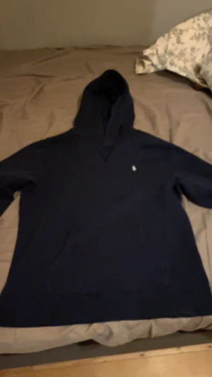 Mörkblå hoodie från Polo Ralph Lauren - Snygg mörkblå hoodie från Polo Ralph Lauren med klassisk gul broderad logga på bröstet. Hoodien har huva och stor känguruficka framtill. Mjuk och skön bomullskvalitet, perfekt för chill dagar. Passar dig som gillar stilren och enkel design. Ca 170 i storlek. Använd kanske fem gånger. Från kids brand store.