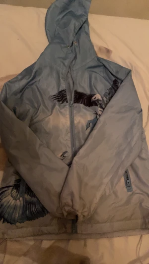 Blå vindjacka från Trapstar med fågeltryck - Säljer en blå vindjacka från Trapstar med snyggt fågeltryck på framsidan. Jackan har huva, dragkedja och två sidofickor med zip. Perfekt för dig som vill sticka ut med streetwear-vibe och cool design.