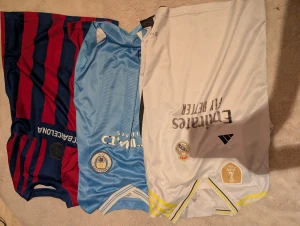 Tre fotbollströjor: Real, City, Barca - Säljer tre snygga fotbollströjor: Real Madrid i vitt med gula detaljer, Manchester City i ljusblått med tunna ränder och FC Barcelona i klassiskt rödblått randigt. Alla har klubbmärken och tryck på bröstet förutom Barcelona, där märket har åkt av. perfekt för dig som älskar fotboll och vill representera ditt lag. Säljer allihopa för ett pris :)
