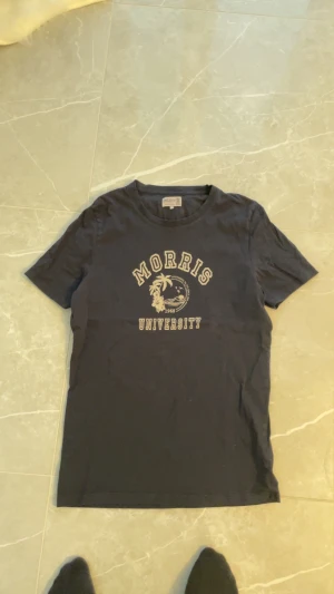 Svart Morris University t-shirt L - Svart t-shirt från Morris i storlek L med klassiskt tryck 'Morris University' och palmmotiv på bröstet. Rund hals och korta ärmar. Skön och mjuk bomullskvalitet som passar perfekt till jeans eller shorts. Snyggt college-inspirerat plagg för en chill look.