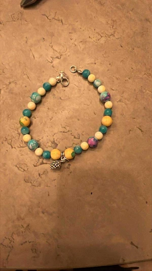 Här har vi har ett armband som jag säljer detta heter ’bluepinkgreenbracelet’ - Namn ”cone-yellow.green/blue artikelnummer ”56Op” 16 cm, Unikt handgjort armband med pärlor i blått, gult, vitt och lila. Armbandet har en silverfärgad charm i form av en liten kotte. Pärlorna är runda och har marmorerade detaljer. Perfekt som personlig accessoar eller present.