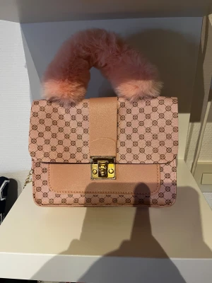 Rosa handväska med päls och kedja - Snygg rosa handväska med mönstrad yta och guldfärgat lås. Väskan har ett fluffigt handtag i rosa päls och en tunn guldfärgad kedja som axelrem. Perfekt för dig som vill sticka ut med en unik accessoar.