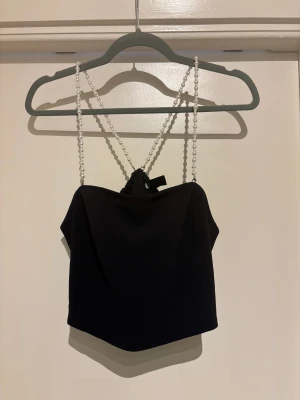 Svart croppad topp med pärlband, Lindex - Tvättråd: 40° Pärlbanden kan tas av, kan ersättas med  andra band. Fästs med krokar.  Köpt november 2024. Nypris 499 kr.  