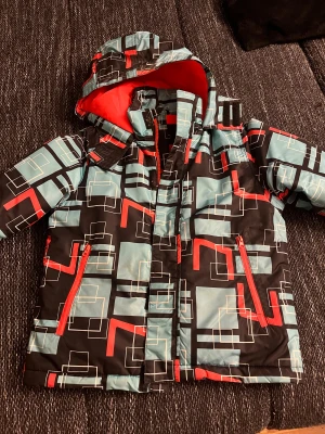 Färgglad pufferjacka med grafiskt mönster - Säljer en pufferjacka med huva och grafiskt mönster i svart, ljusblått och rött. Jackan har röda dragkedjor och ett coolt rutigt mönster med vita linjer. Insidan av huvan är röd och jackan är perfekt för kalla dagar.