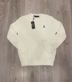 Vit kabelstickad tröja Polo Ralph Lauren - Vit kabelstickad tröja från Polo Ralph Lauren med klassisk rund hals och broderad logga på bröstet. Tröjan har ribbade muddar vid ärmslut och nederkant, vilket ger en snygg och stilren look. Perfekt för dig som gillar tidlös design och premiumkänsla.