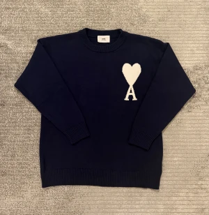 Marinblå stickad tröja från Ami - Snygg marinblå stickad tröja från Ami med stort vitt hjärta och bokstaven A på bröstet. Rund hals och ribbade muddar vid ärmslut och nederkant. Perfekt för dig som gillar stilrena detaljer och vill ha något som sticker ut lite extra.
