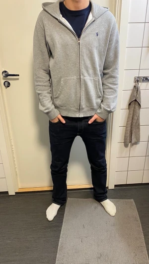 Grå hoodie från Polo Ralph Lauren - Säljer en grå zip-hoodie från Polo Ralph Lauren i storlek XL. Hoodien har huva, dragkedja framtill, två fickor och ett litet blått Polo-logo broderat på bröstet. Mjuk insida i fleece och klassisk passform. Perfekt för chill dagar.