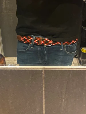 Flätat bälte i svart, rött och vitt - Snyggt flätat bälte med svart bas och inslag av röda och vita detaljer. Bältet har bruna läderdetaljer vid ändarna och ett klassiskt spänne i metall. Perfekt för att ge outfiten lite extra edge och färg. Passar till jeans eller chinos.