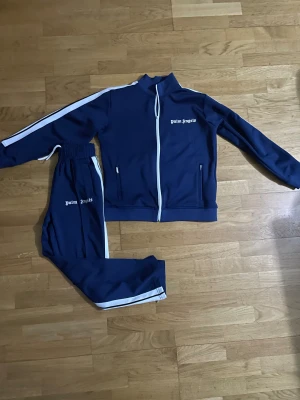 Palm Angels blå tracksuit med vita ränder - Palm Angels tracksuit i marinblått med vita sidoremsor. Setet består av zip-jacka med ståkrage och matchande byxor med elastisk midja. Båda delarna har Palm Angels-logga tryckt i vitt. Materialet är polyester, vilket ger en sportig och skön känsla.